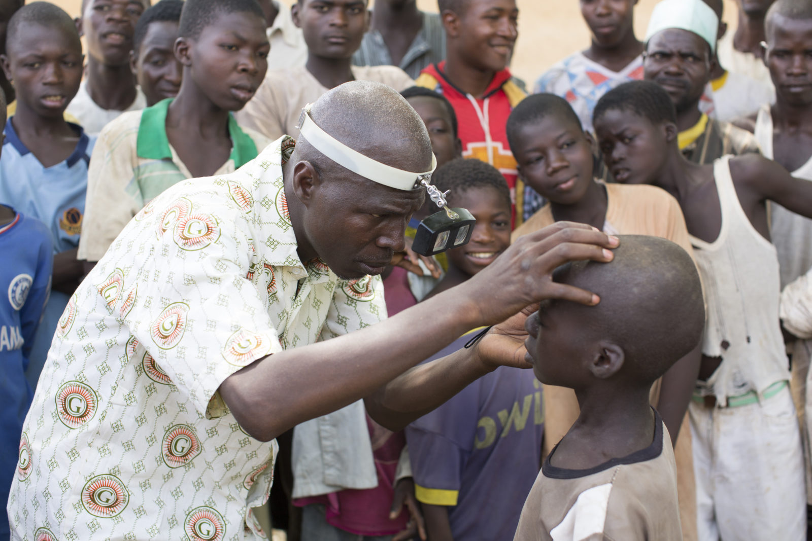 A health worker checks the eyes of a child for signs of disease. Other children are standing behind watching.