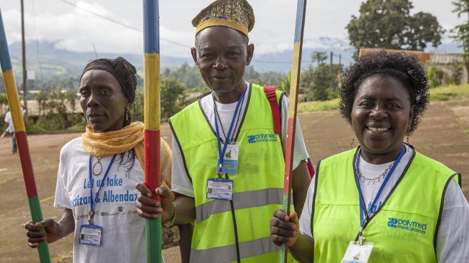 Three health workers stand smiling outside. They are in high-vis jackets and hold dose poles