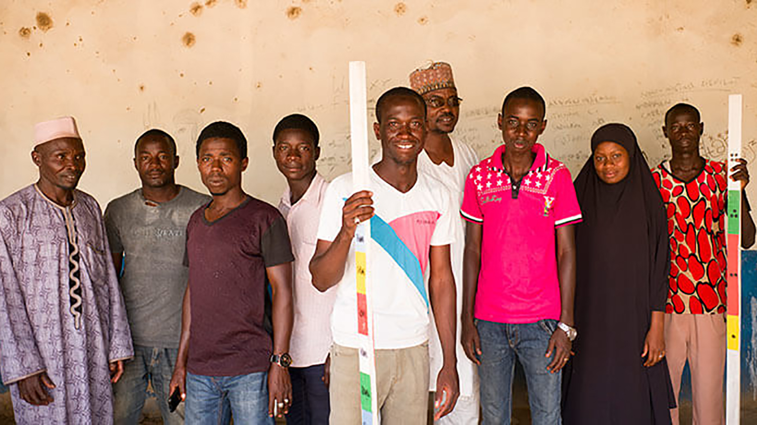 A group of healthworkers stand smiling indoors. One man in the middle holds a measuring stick used for measuring height to allocate the right amount of medication.