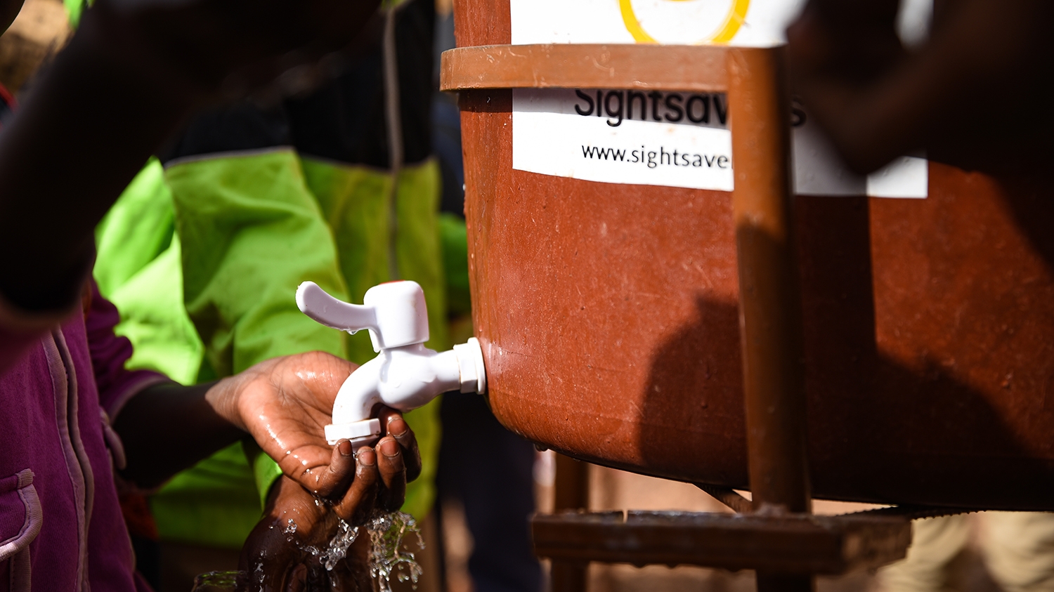 A close-up of someone washing their hands under a tap from a mobile water dispenser. The Sightsavers logo is on the tank.
