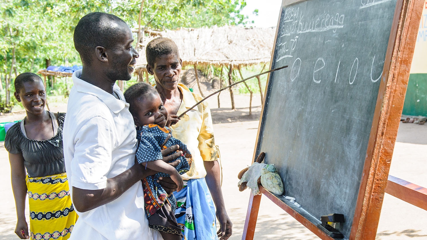 A teacher holds a child up so they can point to something on the blackboard.