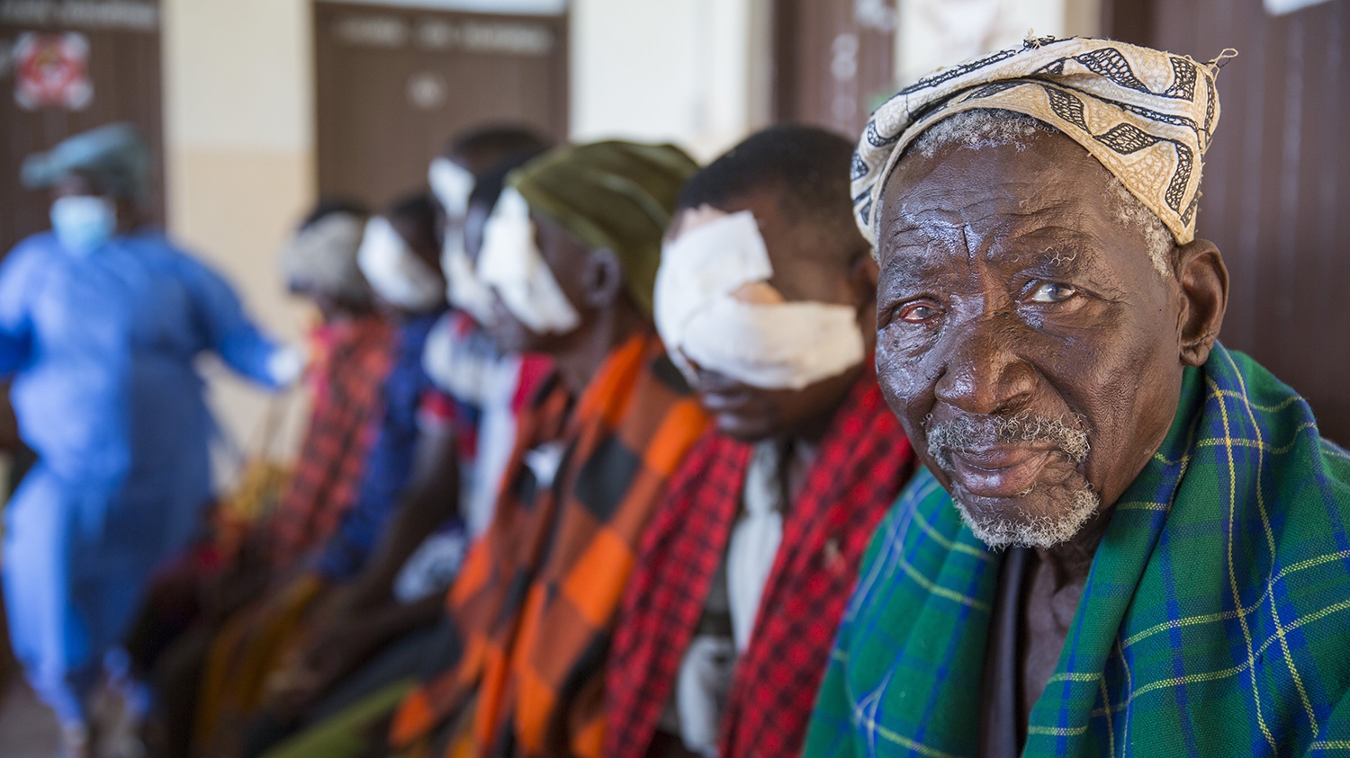 A older man sits in a waiting room alongside other people who have bandages on their eyes after receiving surgery.