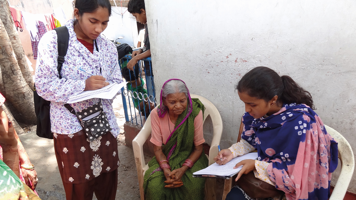 Two female data collectors take information from a woman in Bhopal, India. Two of the women sit on chairs inside and the other stands next to them, writing.