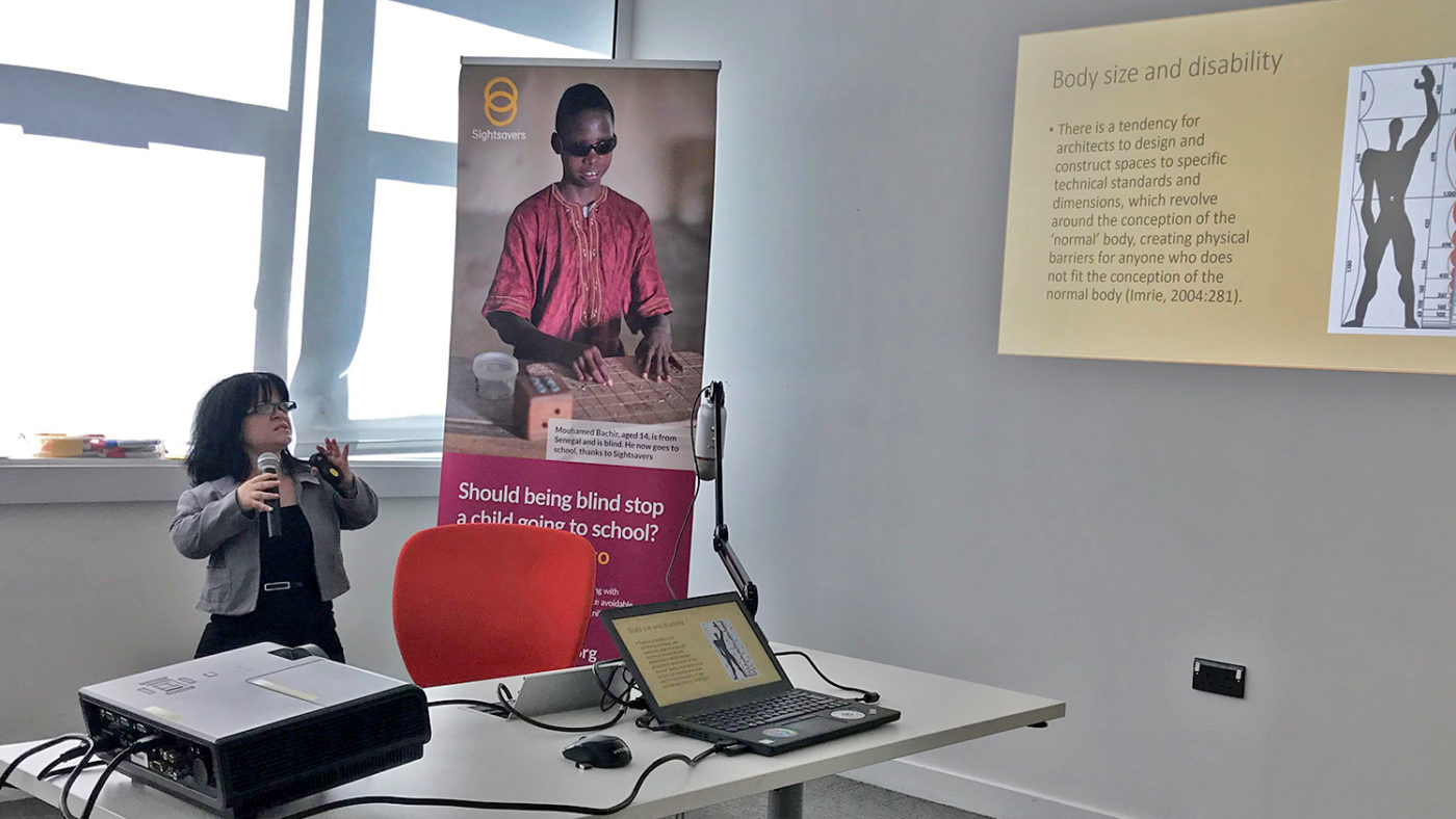 Sightsavers staff member stands behind a desk with a microphone as she gives a presentation about disability in the workplace.