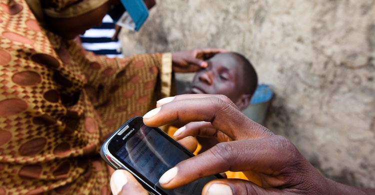 Close up view of a hand holding a phone, in the background a person is having their eye checked as part of the global trachoma mapping project.