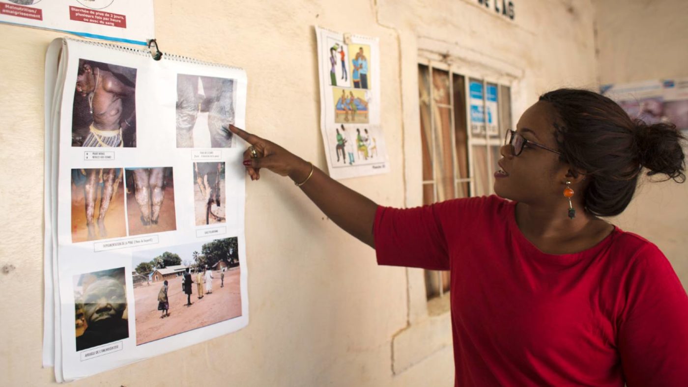 A woman wearing glasses and red t-shirt is pointing at photographs pinned up on the wall. They are images of the symptoms of Lymphatic filariasis.