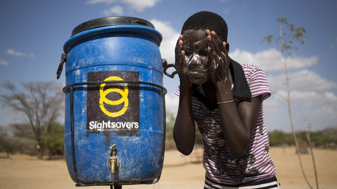 A young boy washes his face outside next to a water container branded with the Sightsavers logo