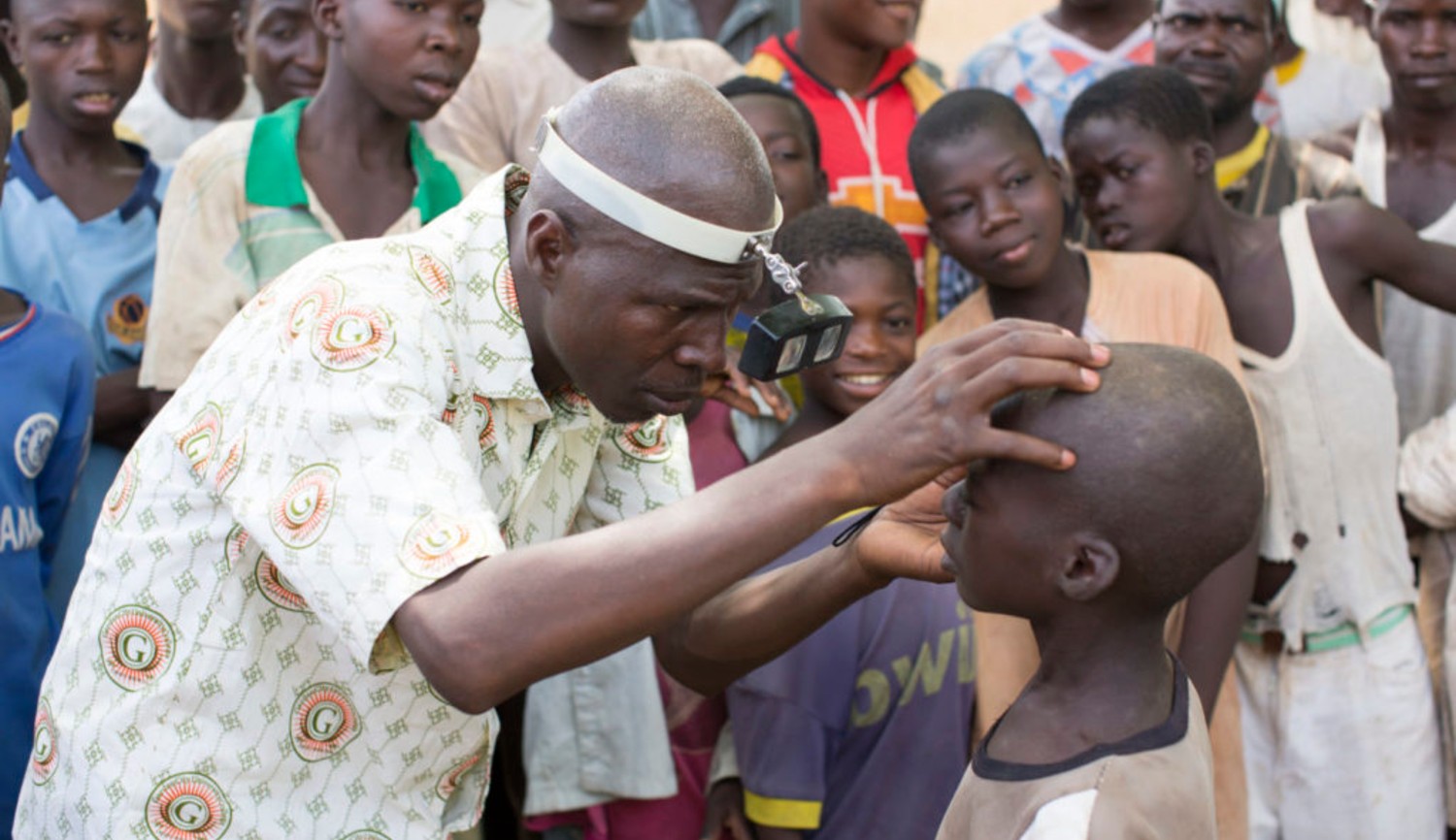 A health worker checks the eyes of a child for signs of disease. Other children are standing behind watching.