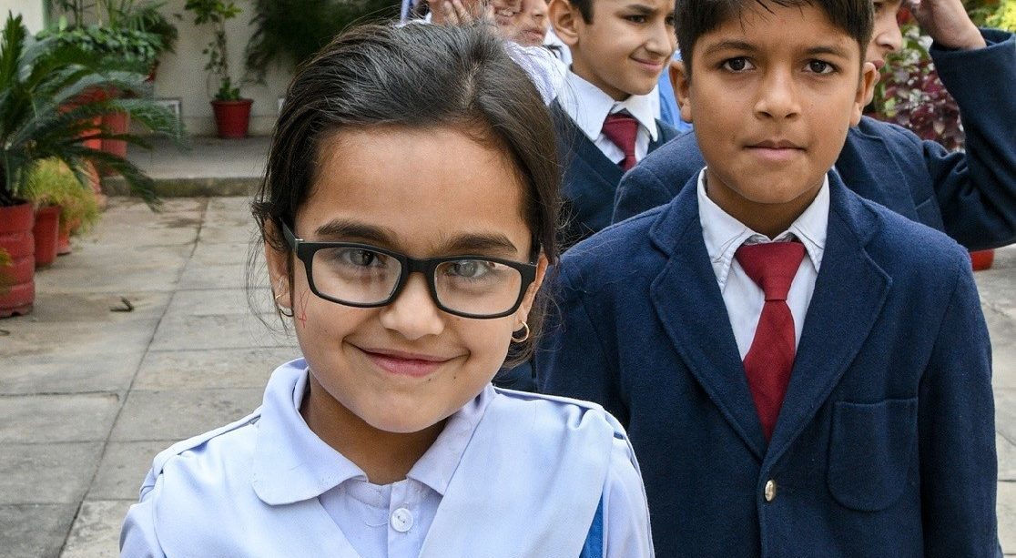 A girl and a boy smile at the camera. The girl is in front and wears glasses.