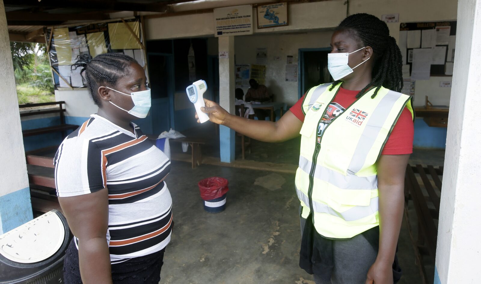 A woman wearing a high-visibility jacket checks the temperature of another woman. Both are wearing masks.