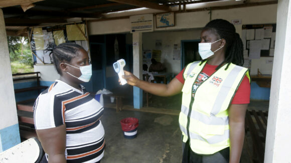 A woman wearing a high-visibility jacket holds up an infrared thermometer and points it at another woman. Both women are wearing facemasks.
