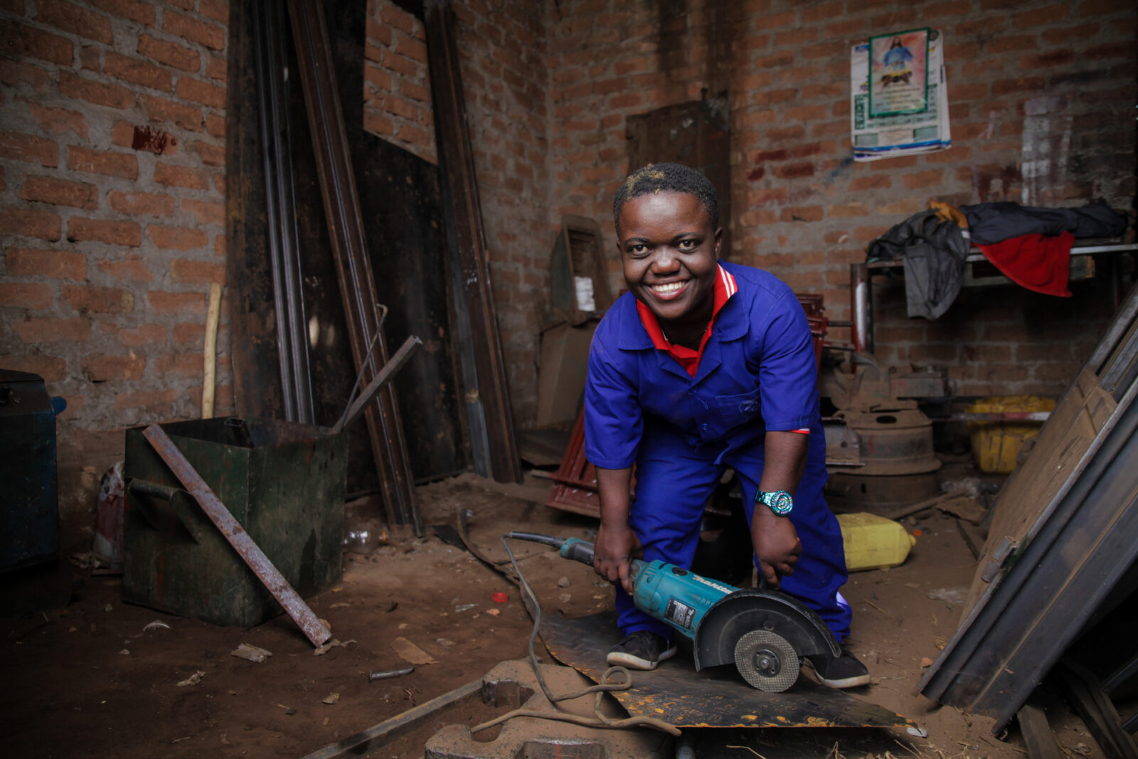 A man with dwarfism is pictured in a workshop, smiling as he uses a saw to cut a piece of metal.