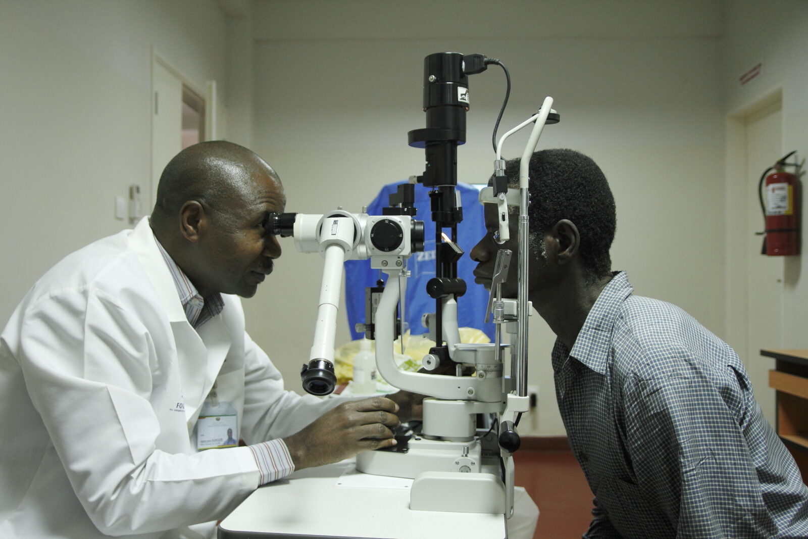A doctor uses a large piece of equipment to test the vision of a man, within a clinic. The man is sitting on a chair, leaning forward and looking into the piece of eye-testing equipment.