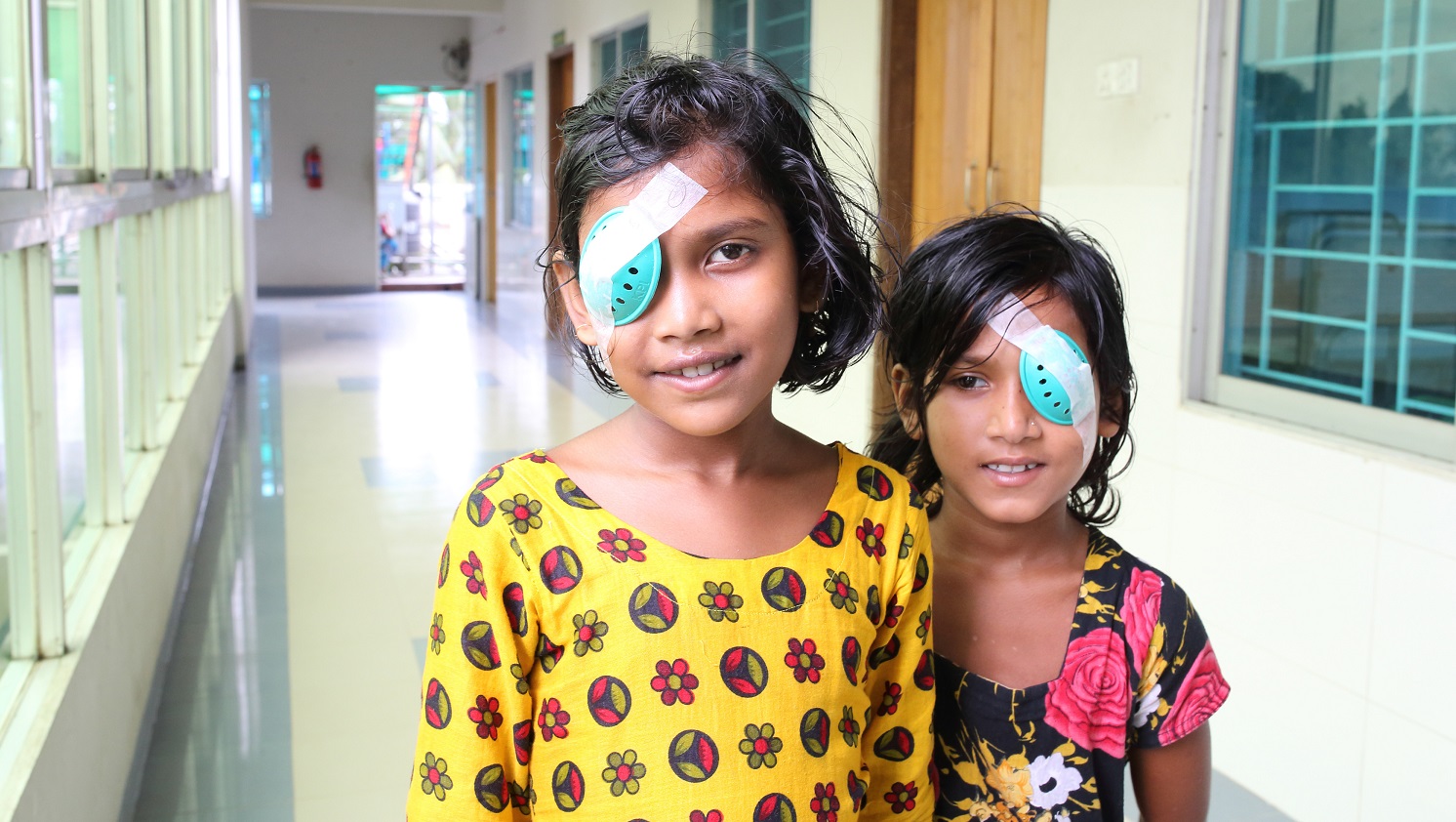 Two young girls stand together in a hospital corridor. Both are wearing colourful clothes, and have smiles on their faces. Both were wearing temporary eye patches after receiving eye surgery.