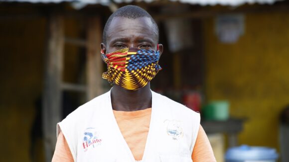 A man wearing a colourful facemask looks into the camera. He is wearing a white uniform that suggests that he is a health worker