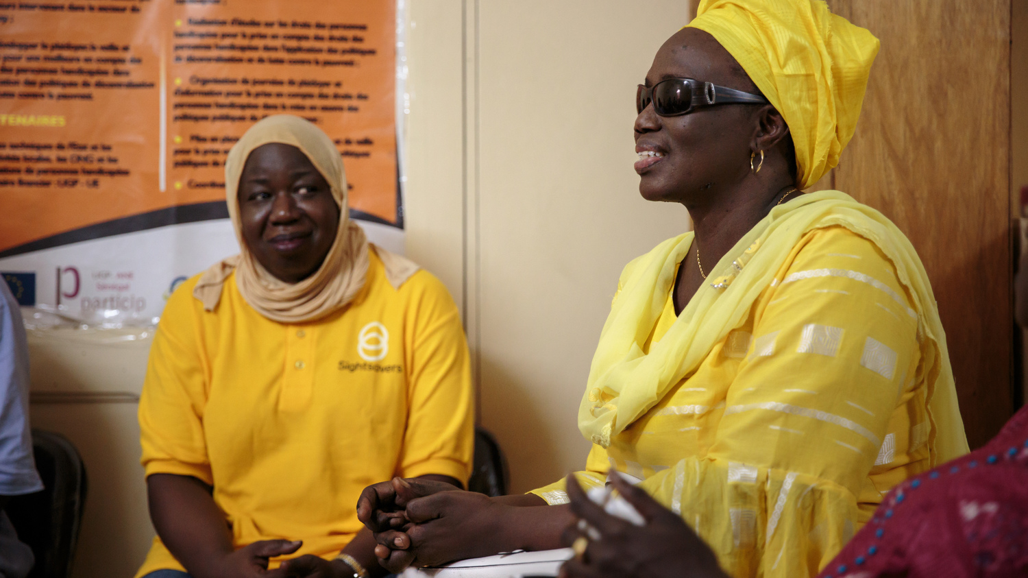 Two women, wearing yellow dresses and headscarves, talk and laugh together.