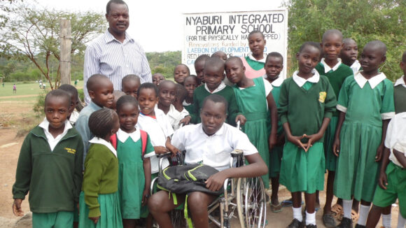 A boy sits in a wheelchair, surrounded by other schoolchildren who are standing up. All of the children are wearing a green school uniform. Behind them is a sign that says, 