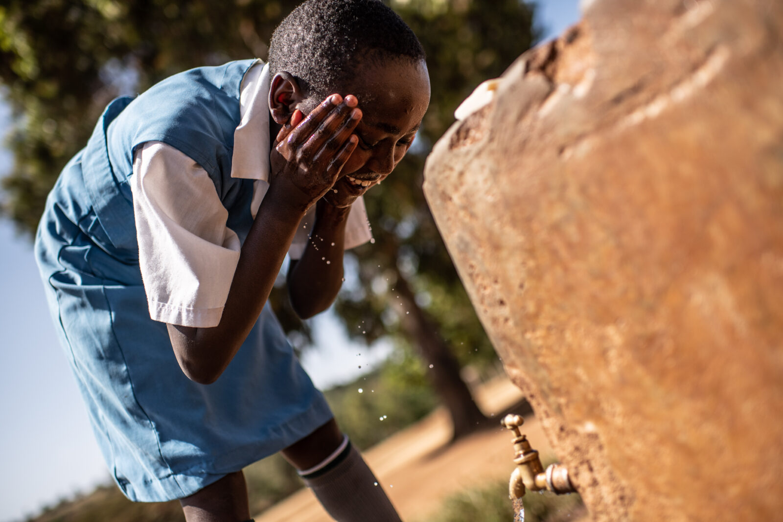 A young girl wearing a blue school uniform smiles while washing her face.