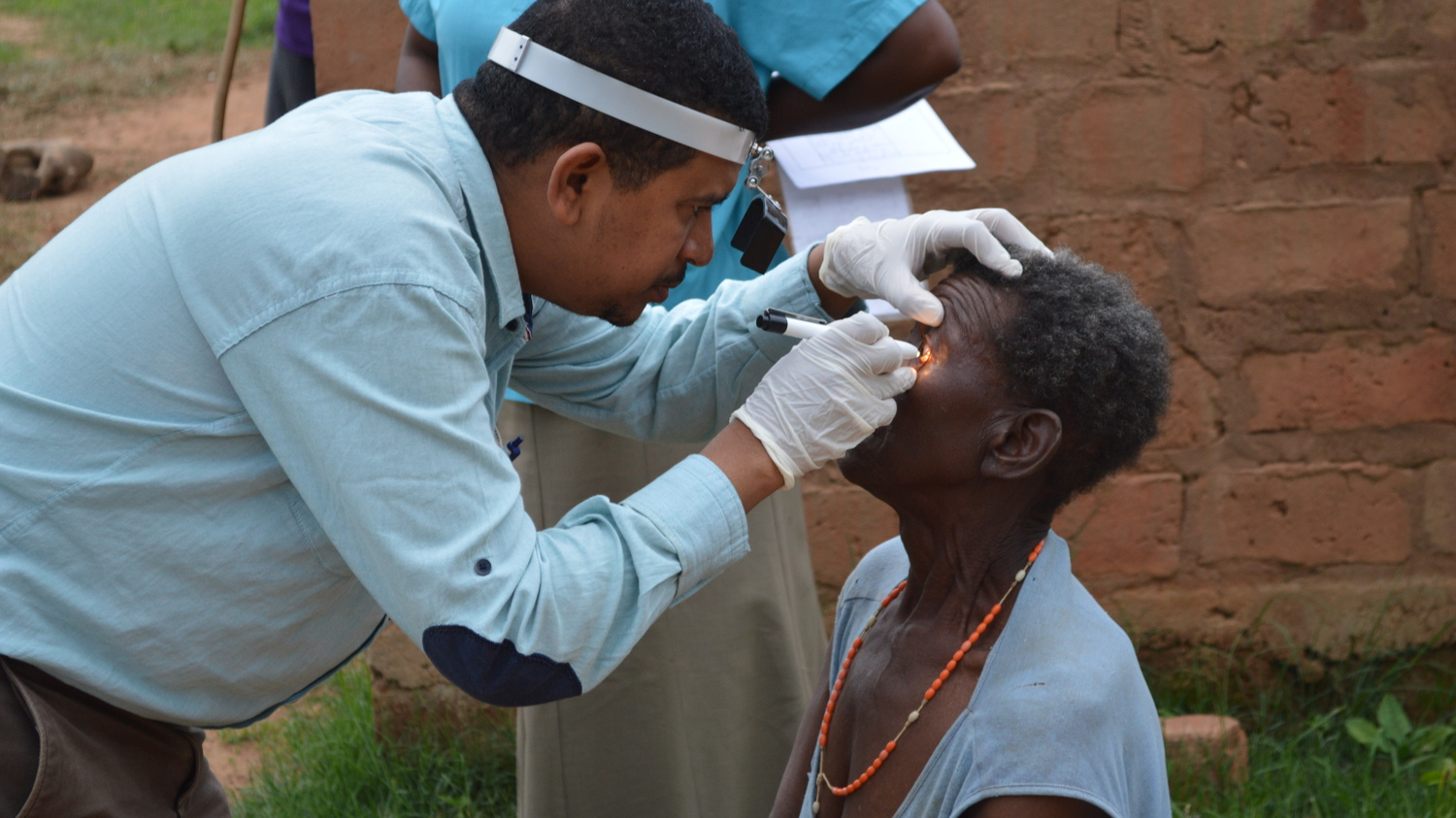 A health professional uses a piece of medical equipment to examine the eye of the patient. The doctor wearing a white shirt, and a female patient is wearing a blue dress and a necklace of beads.