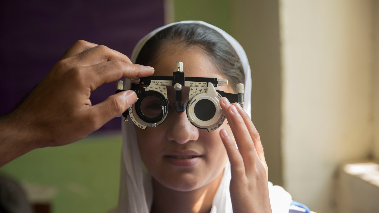 A girl is pictured wearing a white headscarf. She is trying on a special pair of glasses, as part of a vision test. A doctor's hand is seen adjusting the glasses.