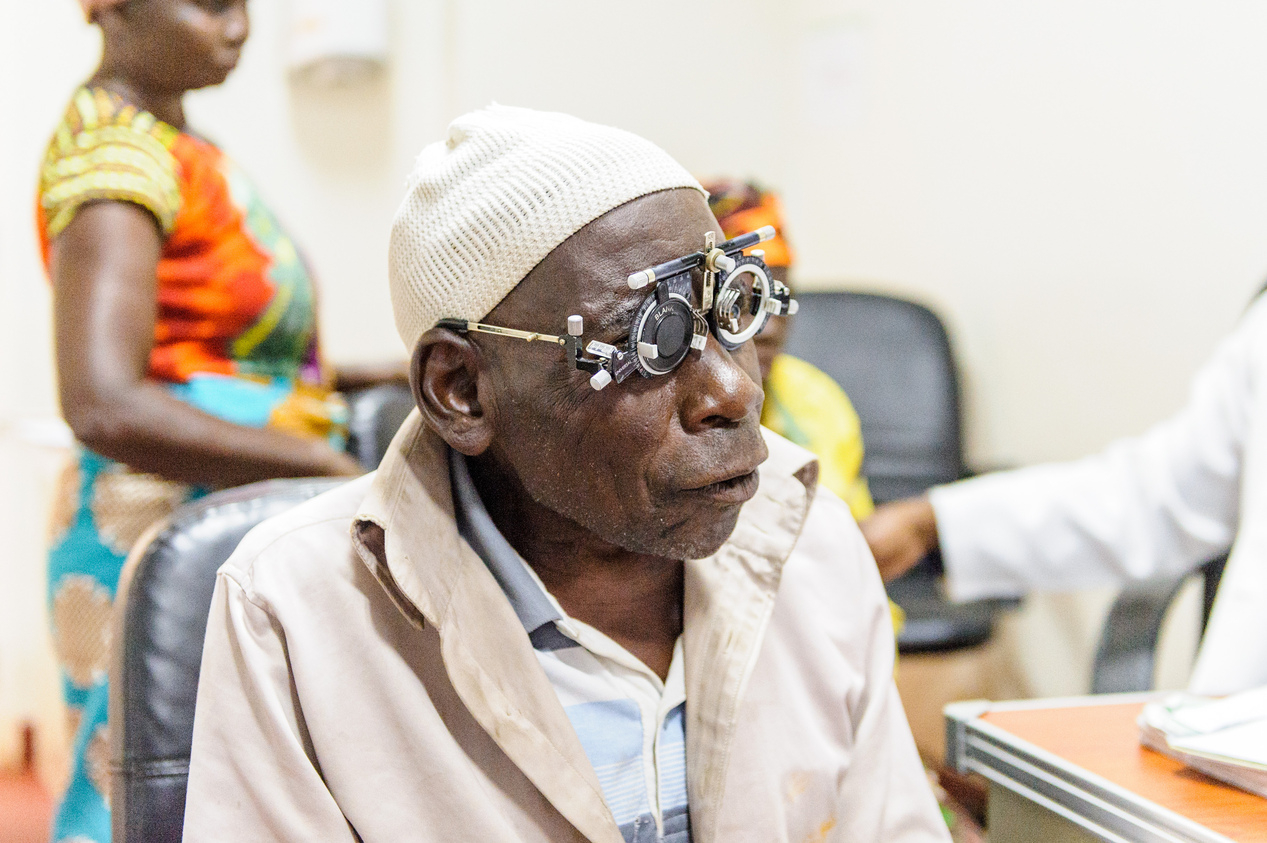 An elderly man, wearing a white jacket and a white cap, sits in a chair. On his face, he is wearing some special glasses that are used for vision screening.