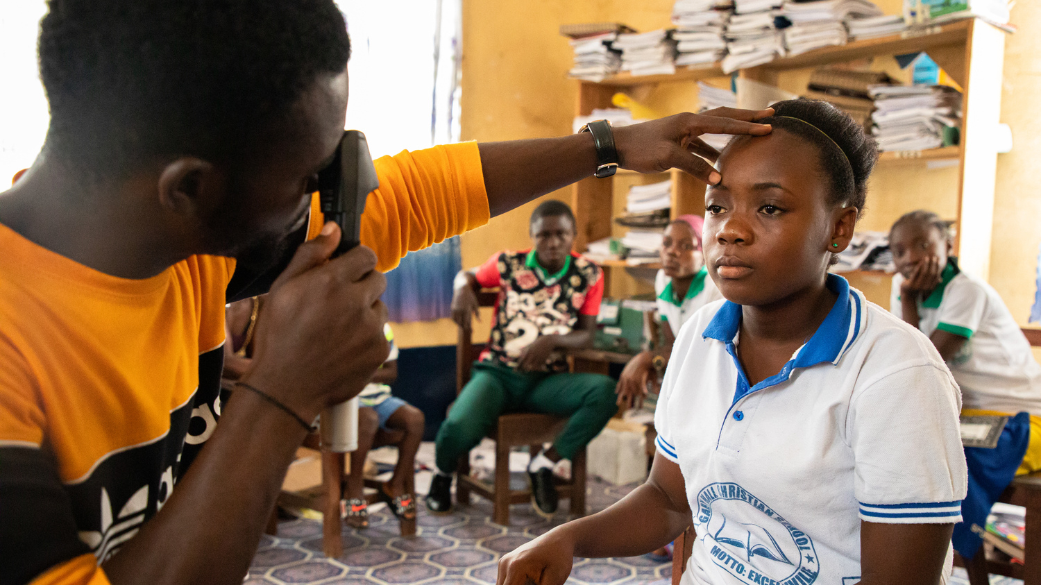 A girl sits on a chair, while a male health worker examines her eyes. The health worker is using a piece of medical equipment with an eye piece and a black handle.