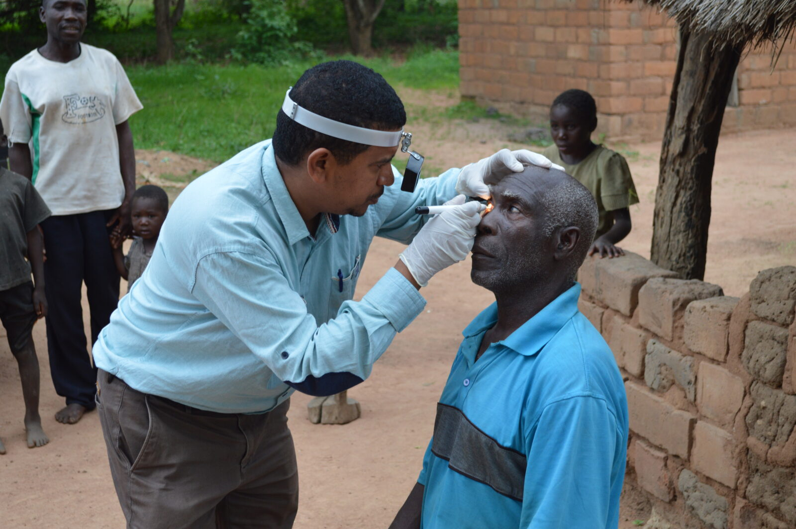 A man wearing a blue shirt and a face visor conducts an eye screening on another man. The man who is receiving the screening is wearing a blue t shirt and sitting down.