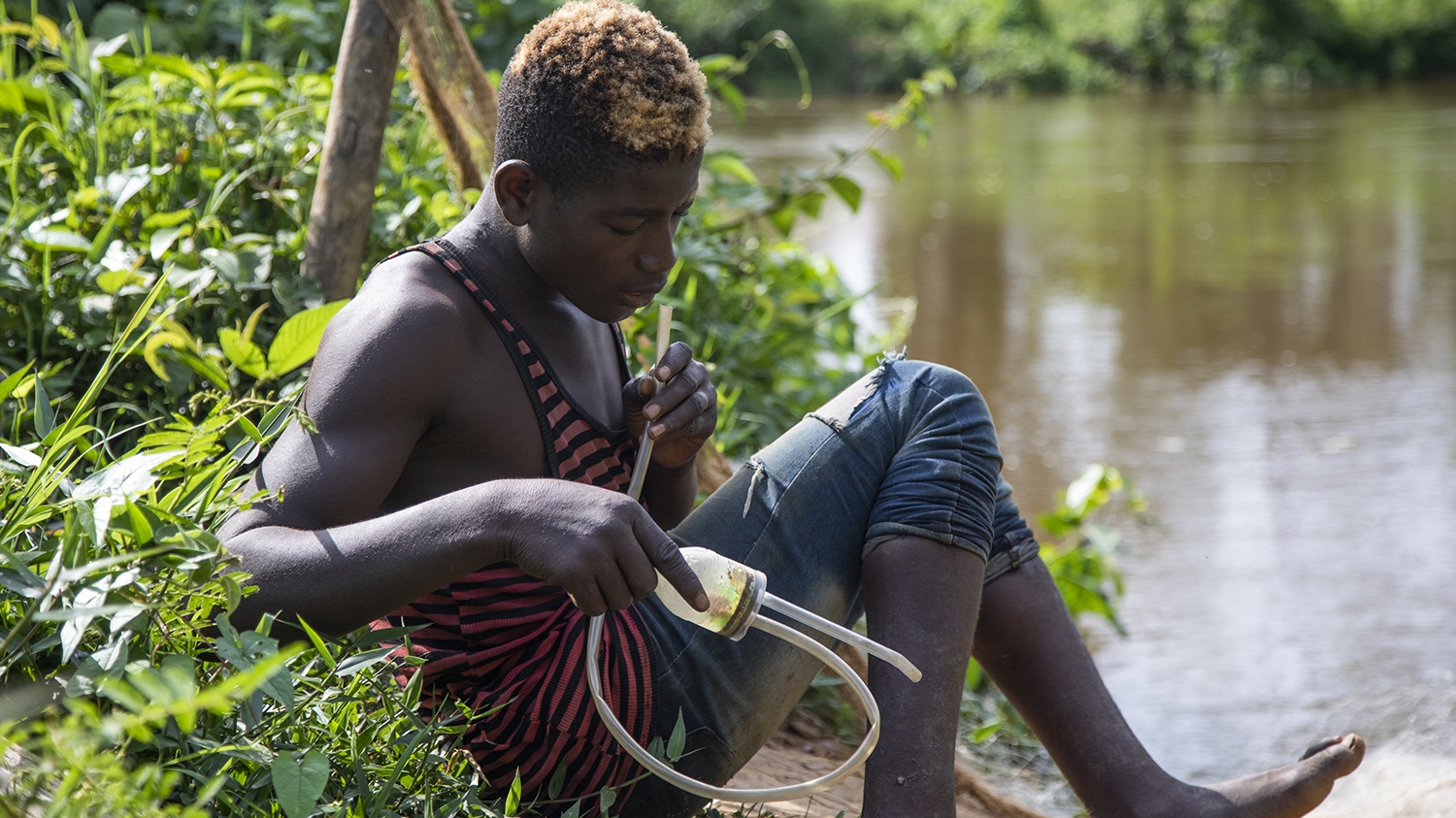 A young man catches black flies on his leg at the edge of a river.