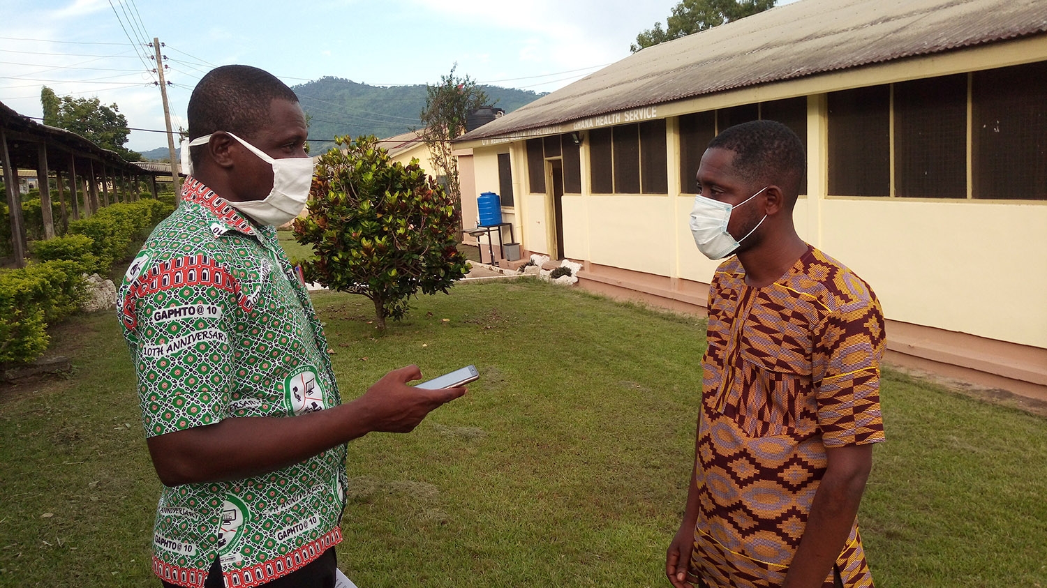 Two men wearing surgical masks stand opposite each other in conversation.