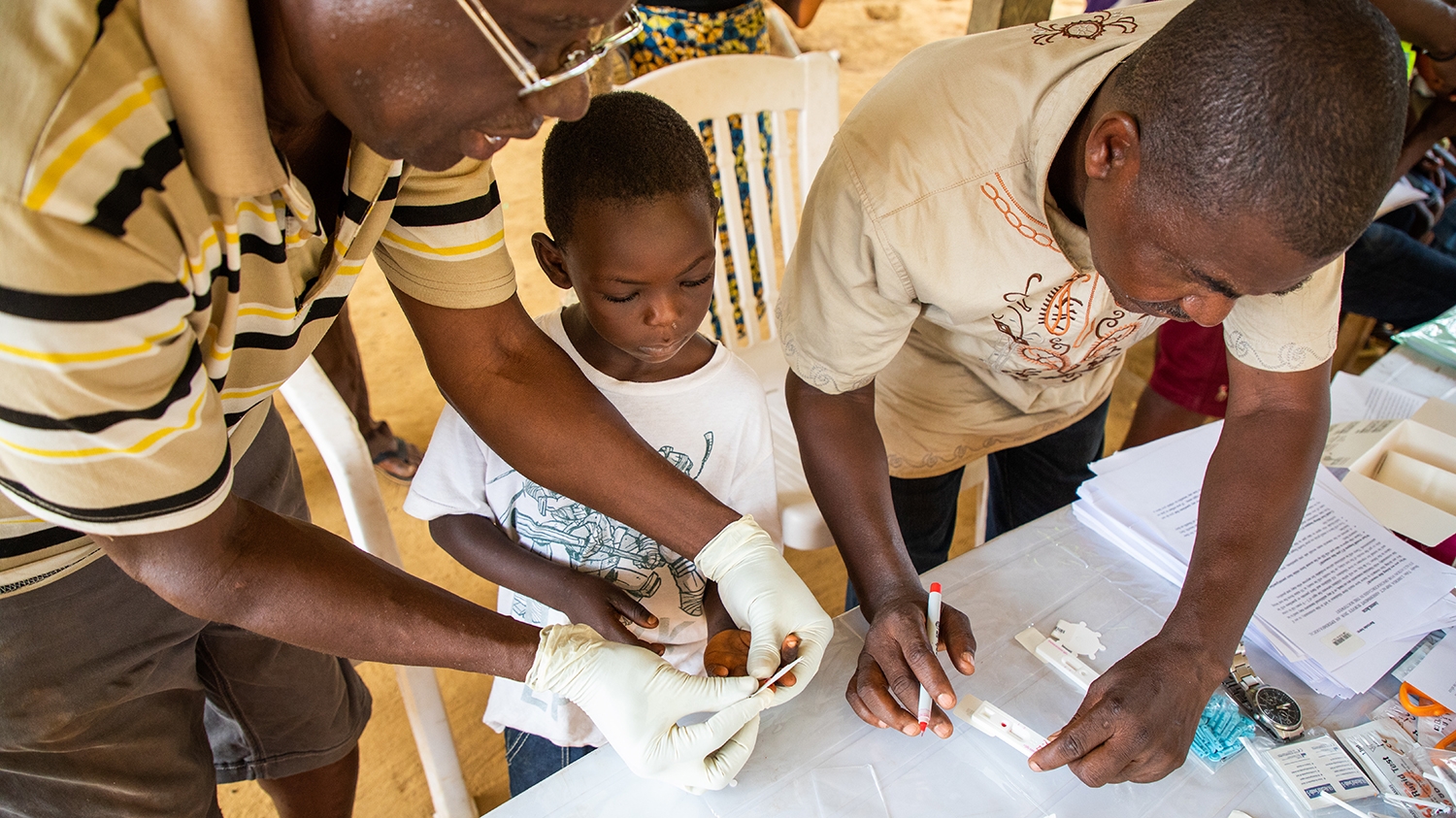 Two male researchers screen a young child from Liberia for onchocerciasis by taking a blood sample.