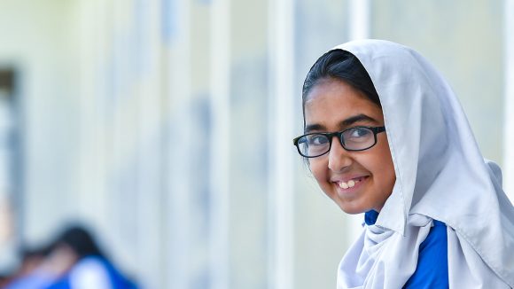 A young girl wearing a head scarf and a pair of glasses smiles as she reads a school textbook.